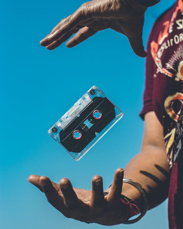 A retro cassette tape floating between hands against a clear blue sky, evoking nostalgia.