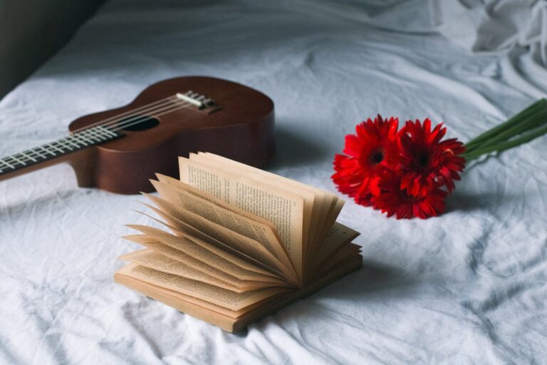 Aesthetic arrangement of an open book, guitar, and red flowers on a bed.